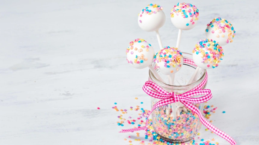 RWK4YG Vanilla cake pops with sprinkles and a cute pink bow over a painted white background.